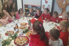 Dzieci siedzą przy wigilijnym stole i częstują się smakołykami i słodkościami.
