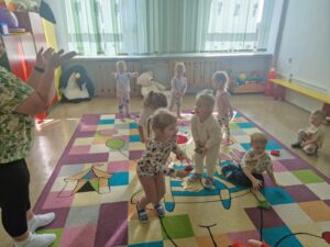 Biedronki wykonują ćwiczenia ruchowe na kolorowym dywanie. Są radosne i roześmiane.