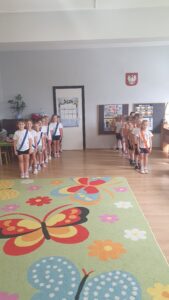 Dzieci z grupy Liski stoją w dwóch rzędach podzielone na drużyny. Przedszkolaki mają szarfy w kolorze niebieskim i  pomarańczowym. Czekają na gwizdek rozpoczynający zawody. 