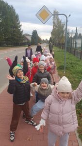 Dzieci z grupy Liski idą chodnikiem. Machają do obiektywu. Są wesołe i uśmiechnięte. Za nimi idą pracownicy przedszkola.