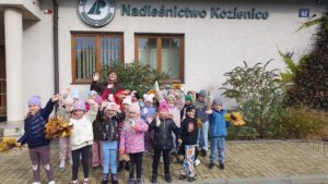 Dzieci wraz z panią leśnik pozują do wspólnego zdjęcia na tle budynku z napisem Nadleśnictwo Kozienice. Machają do obiektywu.