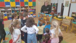 Pani Ola Kołacz prezentuje pracę plastyczną wykonaną przez dzieci, które stoją na dywanie.
