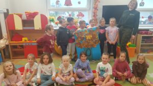 Dzieci z grupy Liski pozują do wspólnego zdjęcia z panią z Kozienickiego Parku Krajobrazowego. Trzymają wspólnie wykonaną pracę plastyczną.