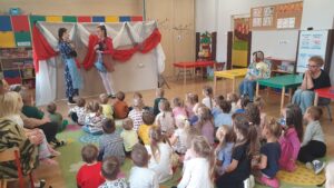 Dzieci siedzą na dywanie w sali przedszkolnej. Oglądają przedstawienie teatralne. Przed nimi występują przebrane aktorki.