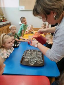 Pani nauczycielka nalewa sok do szklaneczek. Dzieci z niecierpliwością czekają na swoją porcję zdrowia.