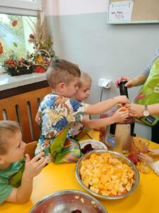 Dzieci wrzucaja pokrojone warzywa i owoce do wyłączonej sokowirówki. Na stole stoi miska z przygotowanymi owocami i warzywami.