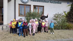Dzieci z grupy Zajączki pozują do wspólnego zdjęcia. Towarzyszy im pani leśnik. Machają do biektywu. W rle widoczny budynek Nadleśnictwa Kozienice.