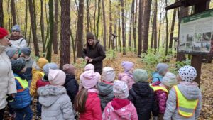 Grupa dzieci słucha prezentacji pani leśnik, dotyczącej ochrony przyrody. Wszyscy znajdują się w lesie.
