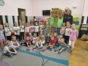 Dzieci z młodszych grup pozują do wspólnego zdjęcia, wraz z panią z Kozienickiego Parku Krajobrazowego. Prezentują wykonaną pracę plastyczną.