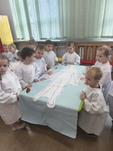Dzieci stoją przy stole na którym leży karton z odrysowanym ciałem człowieka. Widoczne są zdjęcia organów.