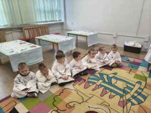 Dzieci siedza na dywanie, ubrane w białe fartuchy. Słuchaja pani prowadzącej warsztaty.
