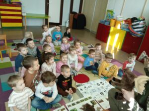 Siedzi siedzą na dywanie i słuchają wykładu pani z Kozienickiego Parku Krajobrazowego. Obok leżą plansze edukacyjne.