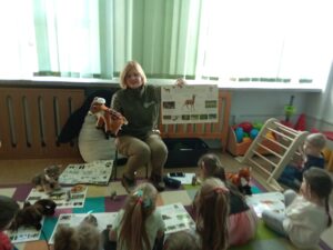 Dzieci siedzą na dywanie a pani z parku krajobrazowego prezentuje zwierzęta na planszy.