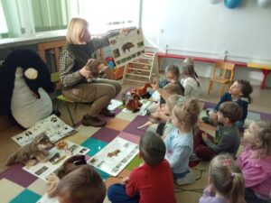 Dzieci siedzą na dywanie i oglądają plansze edukacyjne ukazujące tropy zwierząt leśnych. Pani siedzi na krześle i przedstawia ciekawostki z życia zwierząt.
