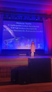 Rozalia Drab recyruje wiersz, stoi na scenie, w tle prezentacja multimedialna z informacją o konkurskie.