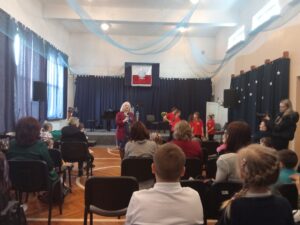 Duża sala w szkole muzycznej. Pani dyrektor zaprasza na koncert. Widzowie siedzą na krzesłach.